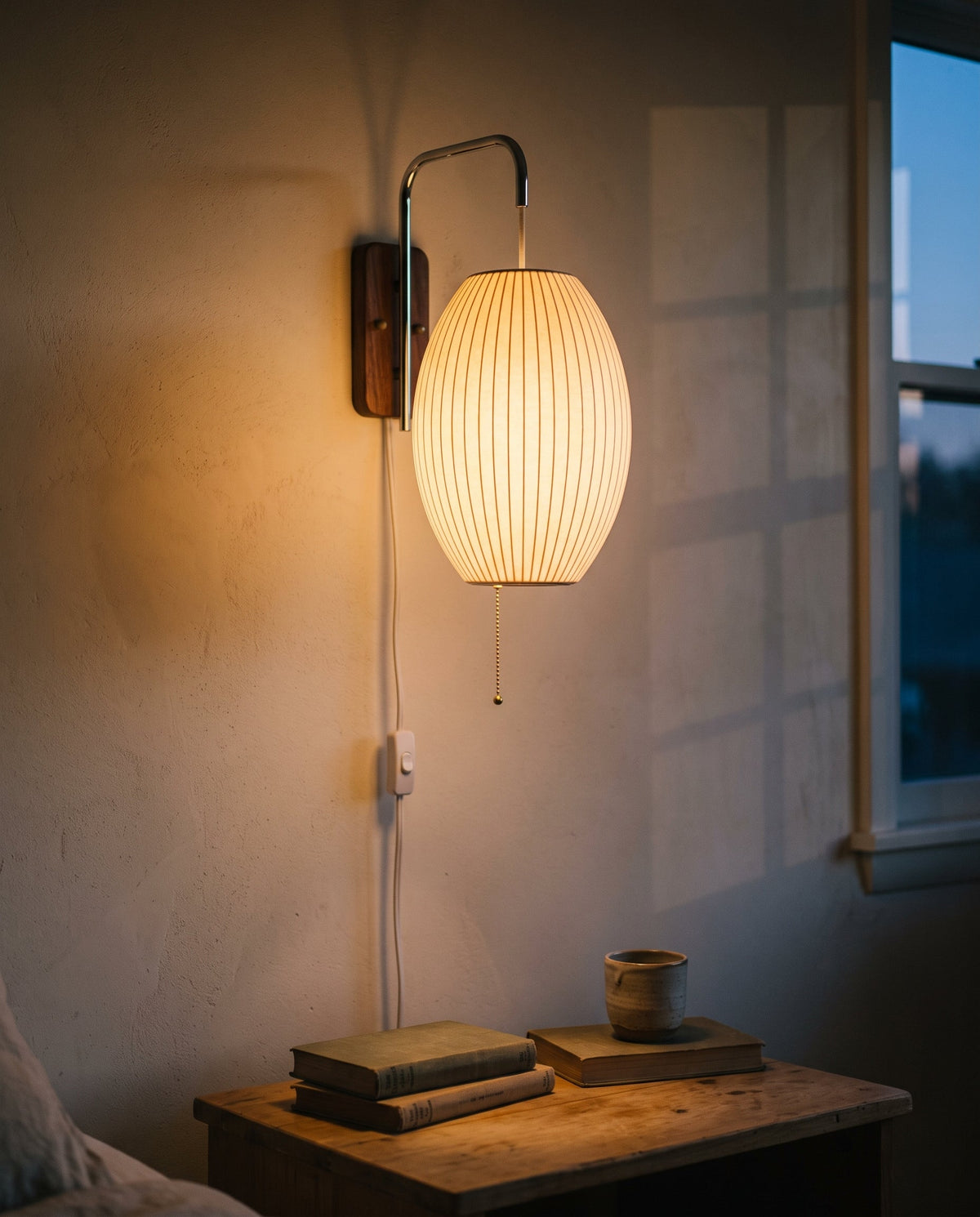 A plug-in wall sconce with an oval, ribbed shade glowing amber at dusk above a wooden bedside table with books