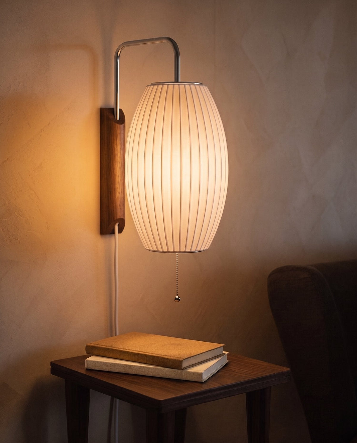 A plug-in wall sconce with a ribbed barrel-shaped shade glowing amber at dusk above a small wooden table with two plain books