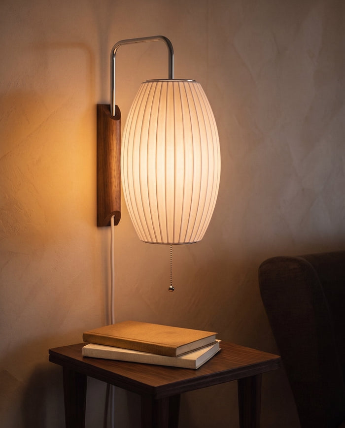 A plug-in wall sconce with a ribbed barrel-shaped shade glowing amber at dusk above a small wooden table with two plain books