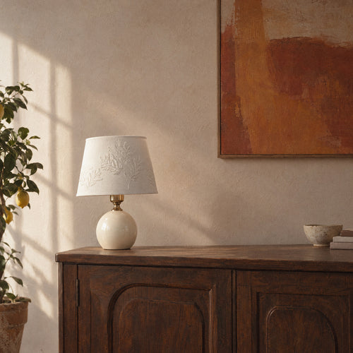 Cream ceramic table lamp with a white leaf-embroidered shade on a walnut sideboard in warm window light