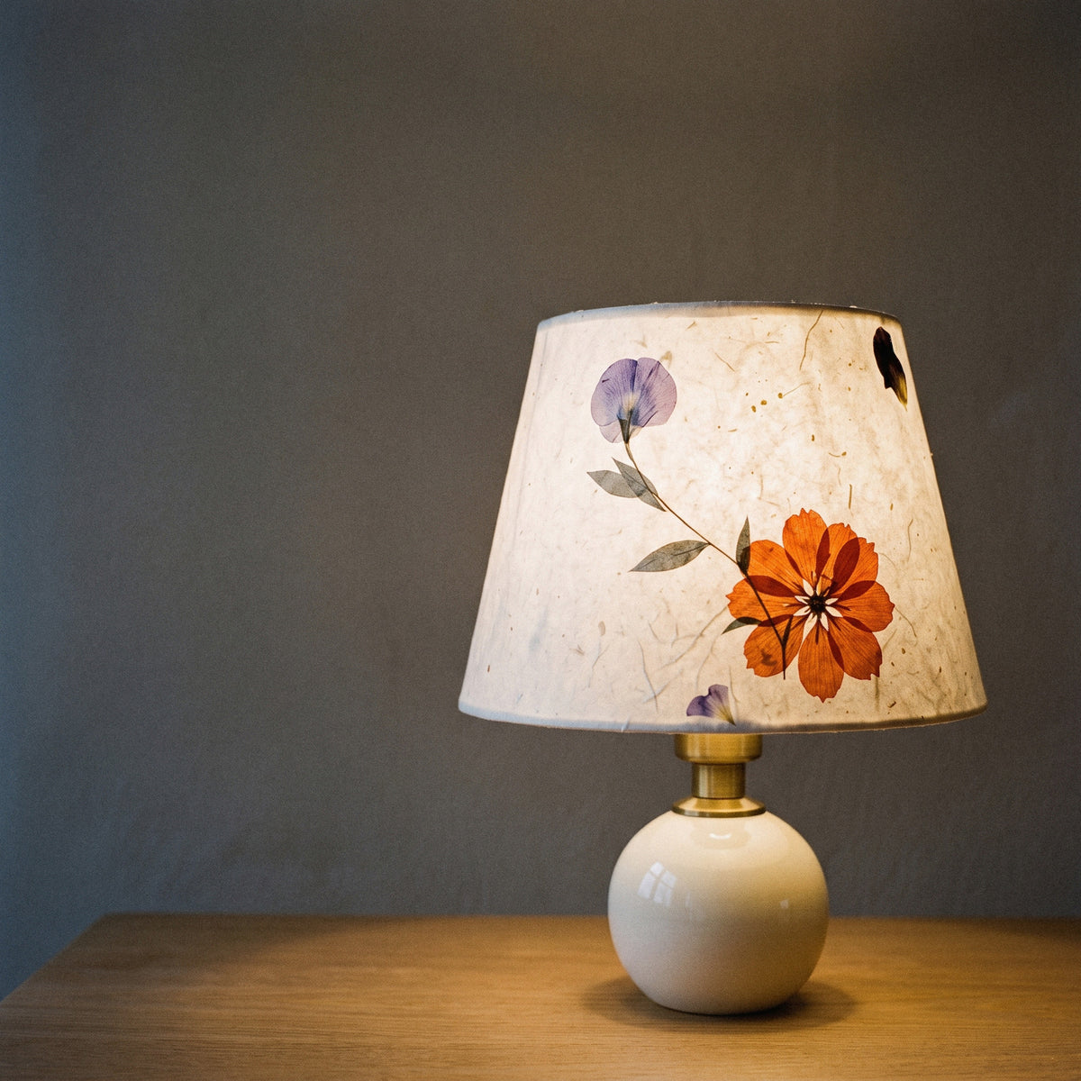Cream ceramic table lamp with a pressed-flower paper shade, lit at dusk with the petals glowing