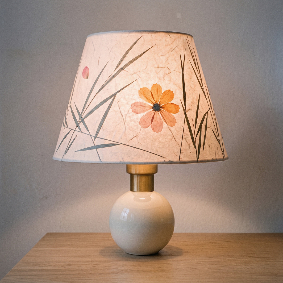 Cream ceramic table lamp with a pressed-flower and grass paper shade, lit at dusk with a soft peach glow