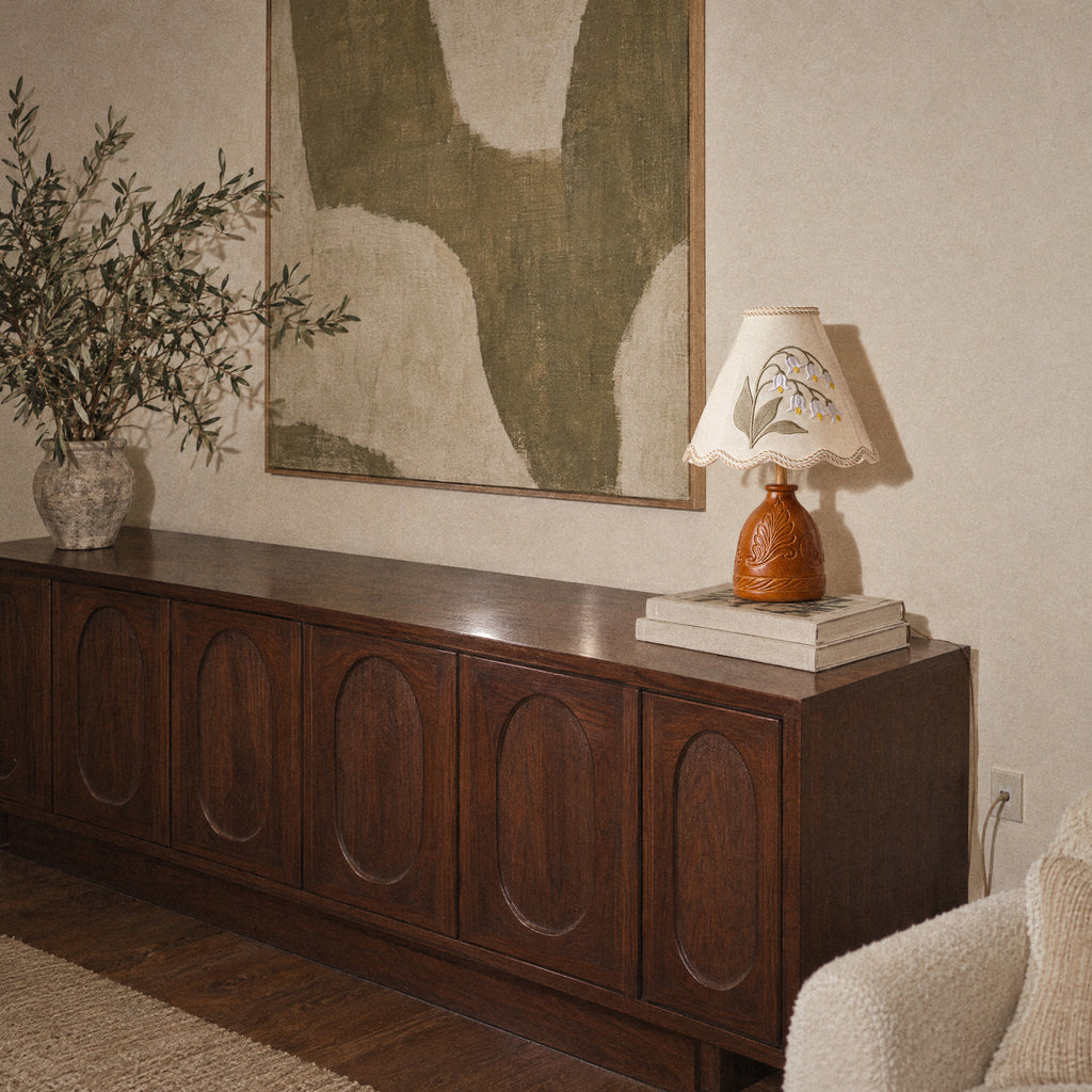 Lily of the Valley table lamp on a walnut sideboard with an olive plant and a canvas, wide room view