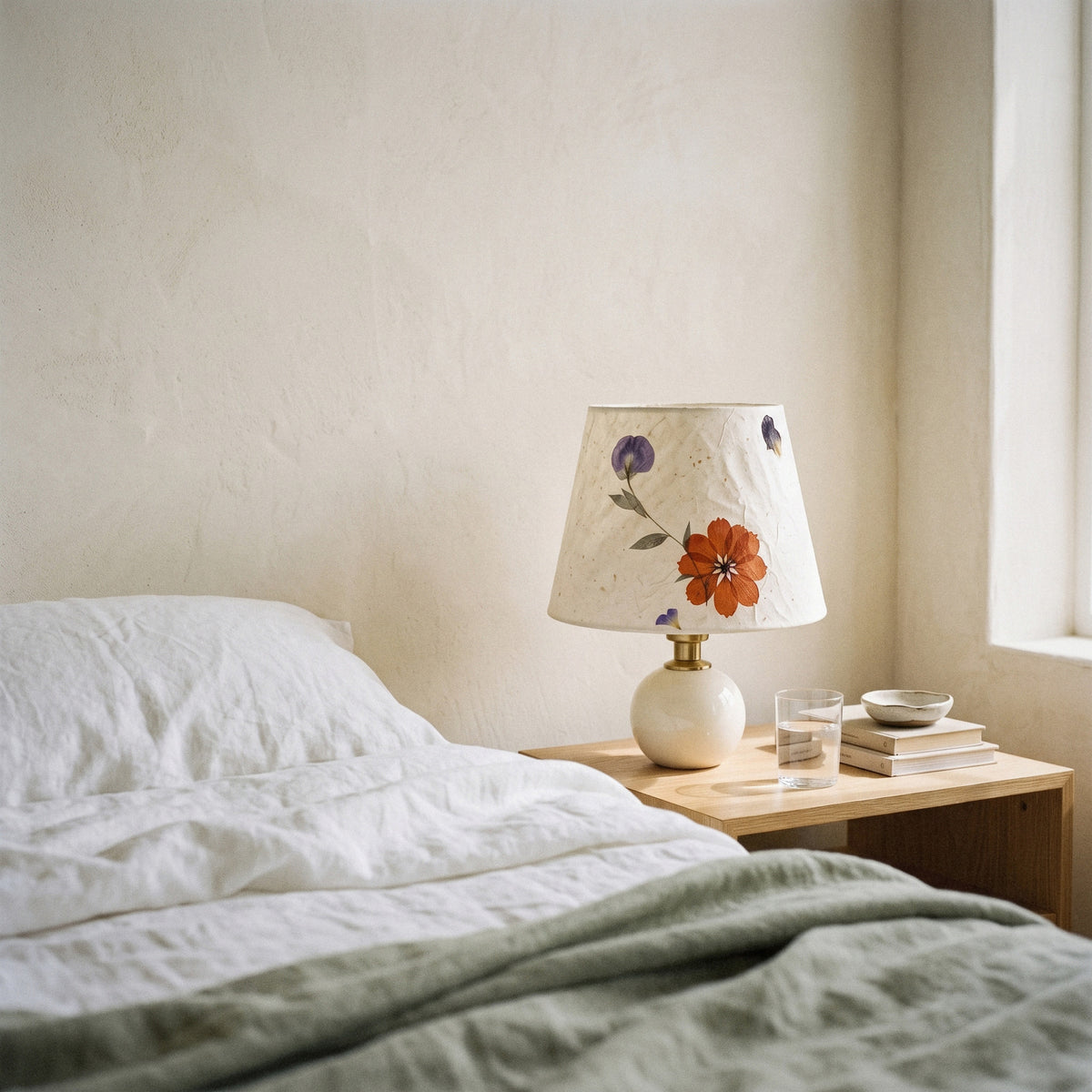 Cream ceramic table lamp with a pressed-flower paper shade on a bedside table with linen