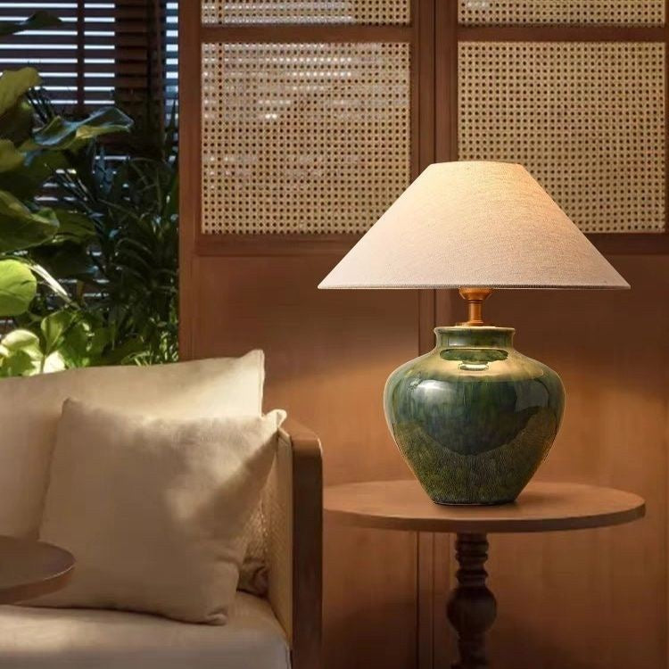 Green ceramic table lamp switched on at night on a side table, casting warm light against a cane screen.