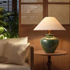 Green ceramic table lamp switched on at night on a side table, casting warm light against a cane screen.