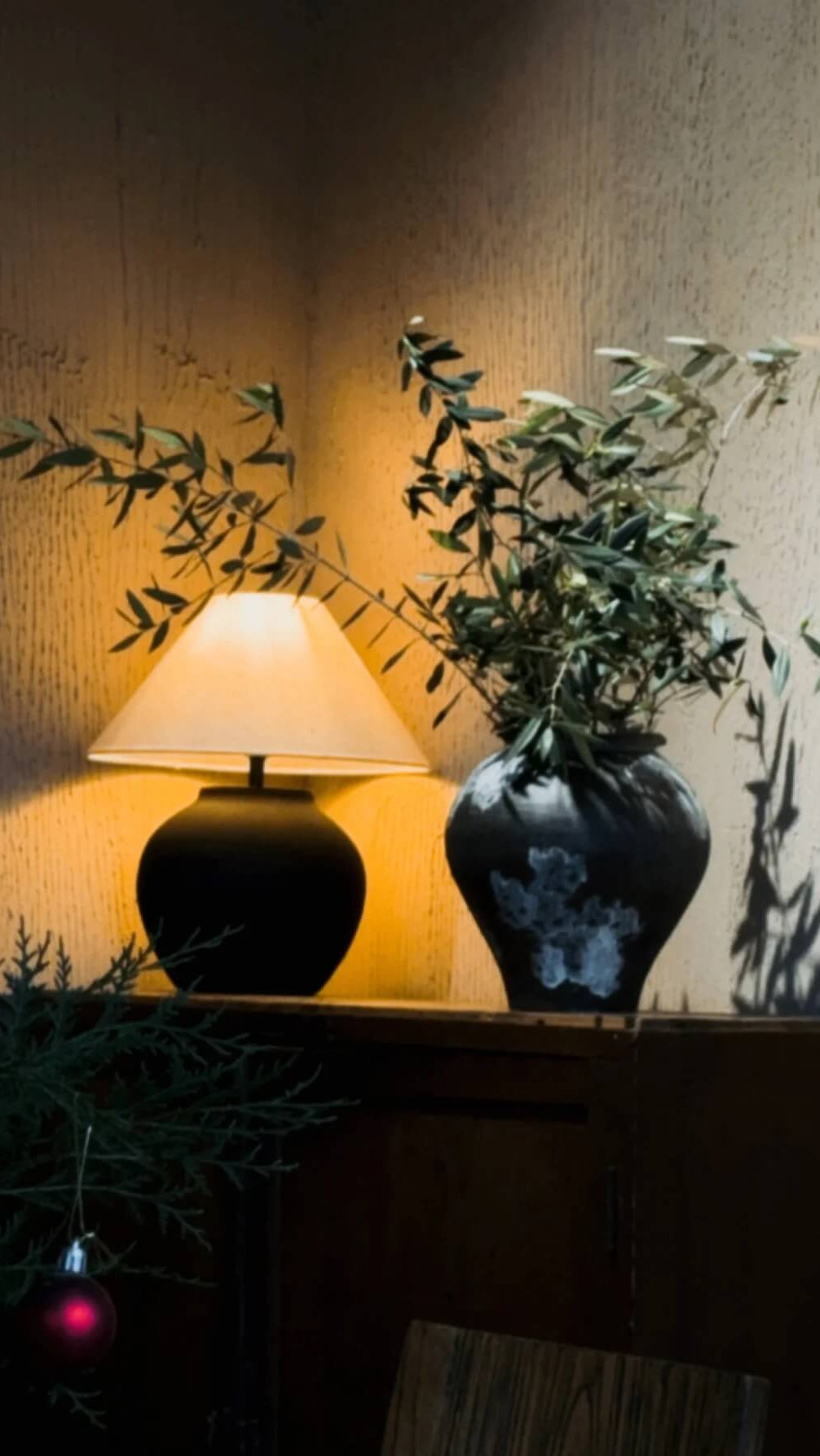 Lamp and vase on a surface with plants and a textured wall in the background