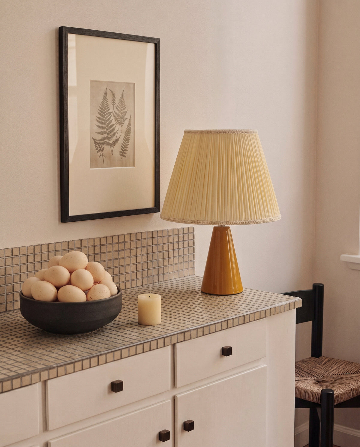 Amber glazed ceramic table lamp with a cream pleated shade on a tiled counter beside a bowl of eggs and a small candle, with a framed fern print on the wall