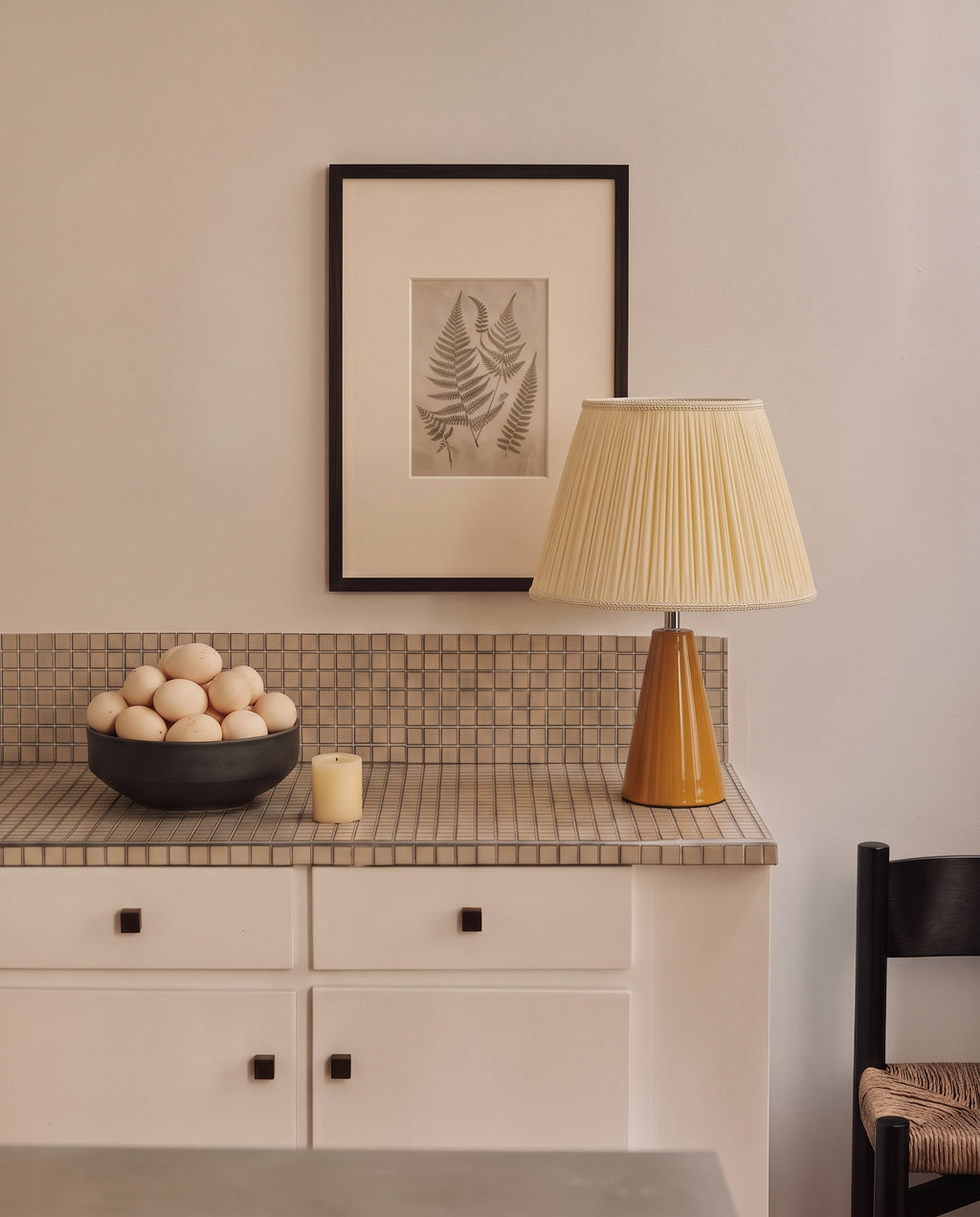 Amber glazed ceramic table lamp with a cream pleated shade on a tiled kitchen counter, beside a bowl of eggs and a small candle