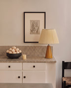 Amber glazed ceramic table lamp with a cream pleated shade on a tiled kitchen counter, beside a bowl of eggs and a small candle