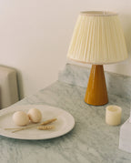 Amber glazed ceramic table lamp with a cream pleated shade on a grey-veined marble counter, beside a plate with two eggs and wheat stems and a cream pillar candle