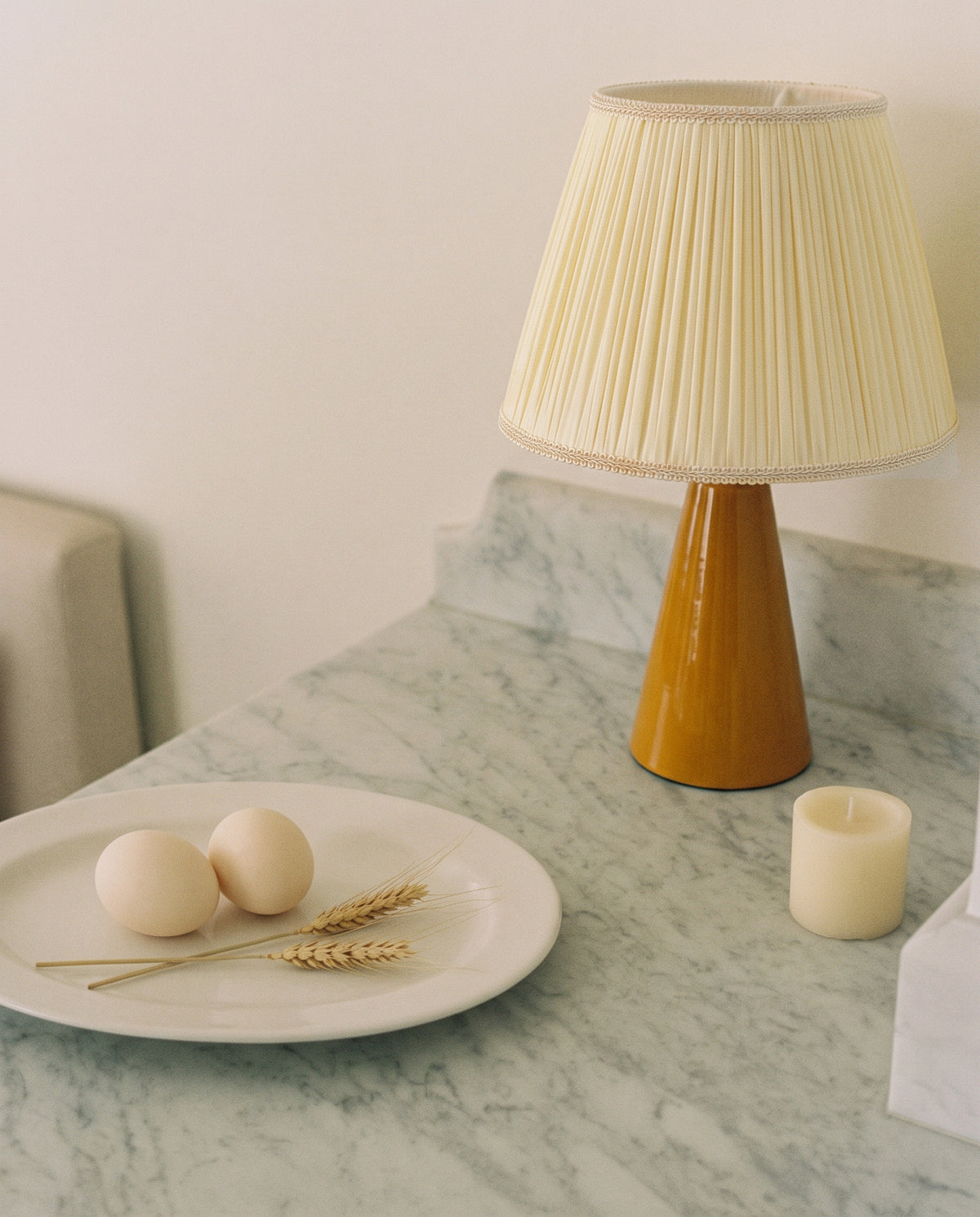 Amber glazed ceramic table lamp with a cream pleated shade on a grey-veined marble counter, beside a plate with two eggs and wheat stems and a cream pillar candle
