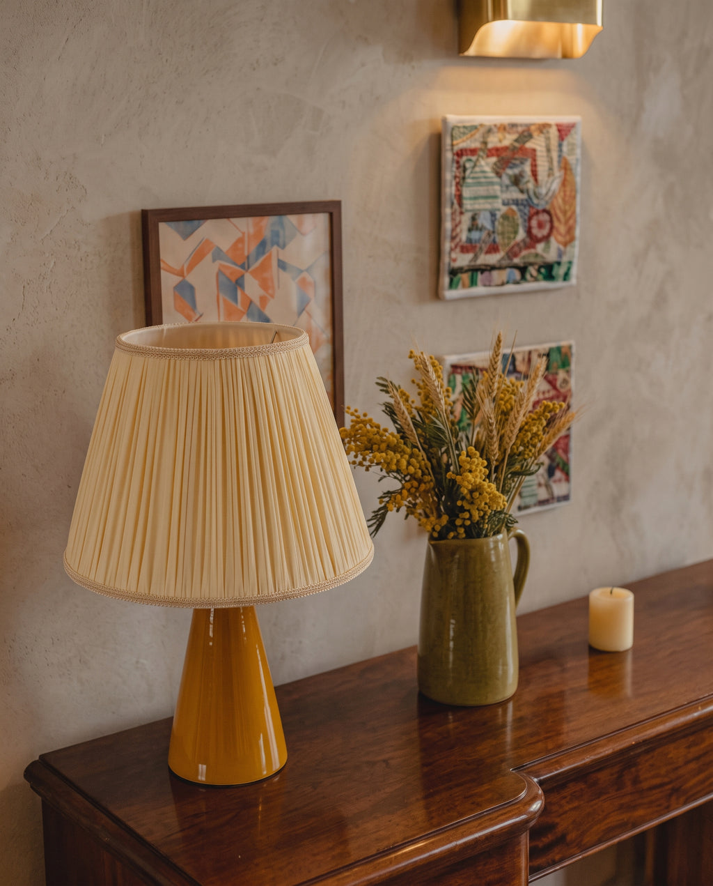 Amber glazed ceramic table lamp with a cream pleated shade on a dark wooden console, beside a green jug of dried mimosa and wheat, with framed textile art on a plaster wall