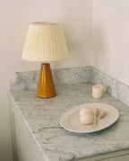 Amber glazed ceramic table lamp with a cream pleated shade on a marble counter, seen from the left side