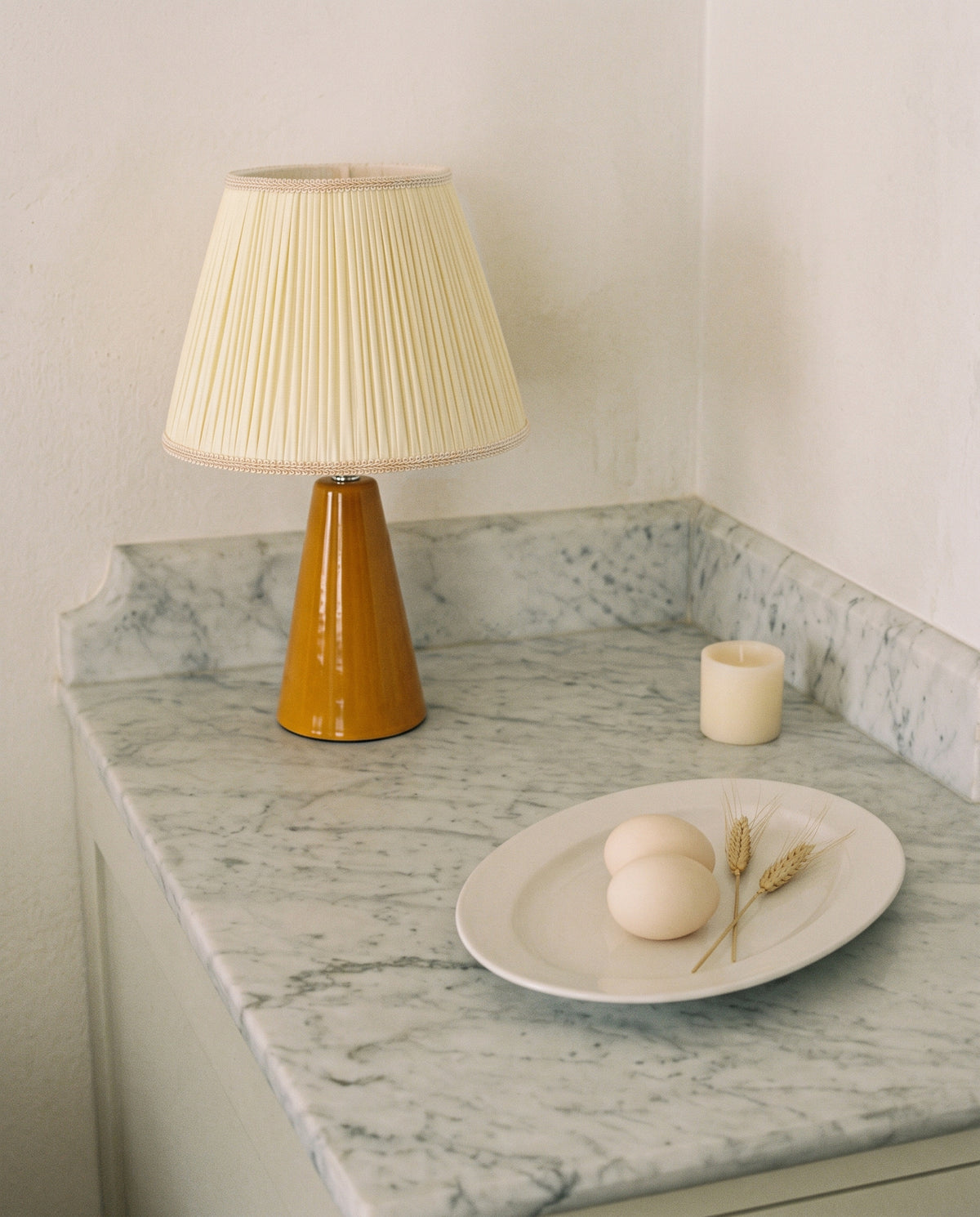 Amber glazed ceramic table lamp with a cream pleated shade on a marble counter, seen from the left side