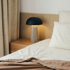 Black dome bedside table lamp with a striped ceramic base on a light wood nightstand beside a linen bed