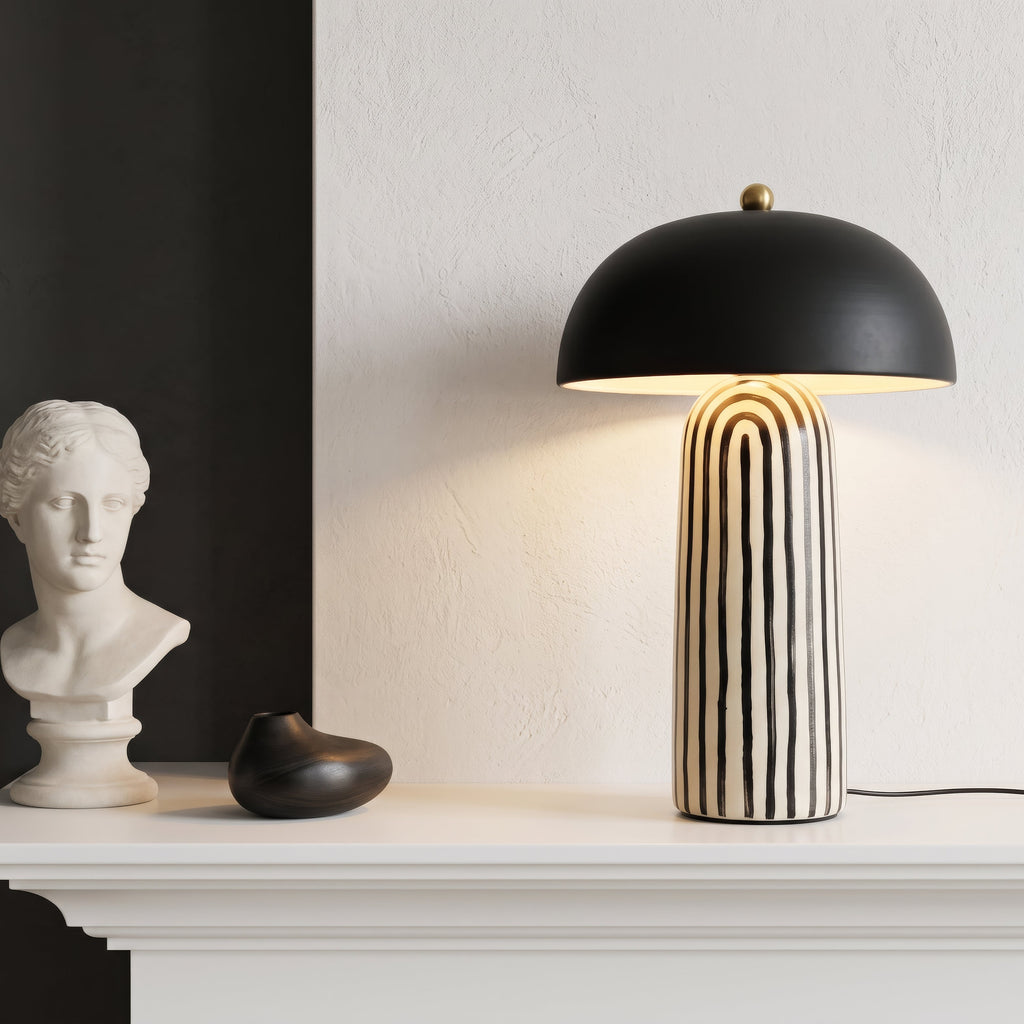 A modern table lamp with a matte black dome shade and a cream-and-black vertically striped ceramic base, styled on a white mantelpiece next to a white classical bust sculpture.