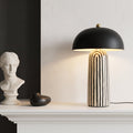 A modern table lamp with a matte black dome shade and a cream-and-black vertically striped ceramic base, styled on a white mantelpiece next to a white classical bust sculpture.