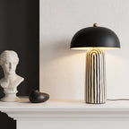 A modern table lamp with a matte black dome shade and a cream-and-black vertically striped ceramic base, styled on a white mantelpiece next to a white classical bust sculpture.