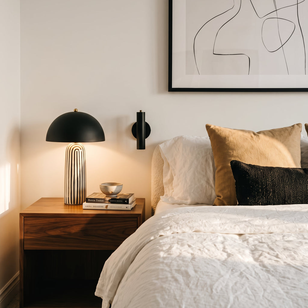 A modern bedroom scene featuring a wooden nightstand with a black dome lamp and books, next to a bed with white textured bedding, a gold pillow, and a black textured pillow. A minimalist black line art print hangs on the wall above.