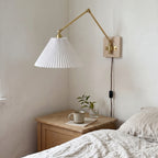Plug-in swing arm wall sconce with a pleated shade and brass arm, mounted above a bedside table next to a linen bed