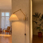 Plug-in swing arm wall sconce with a lit pleated shade on a plaster wall in a sunlit room with herringbone floors