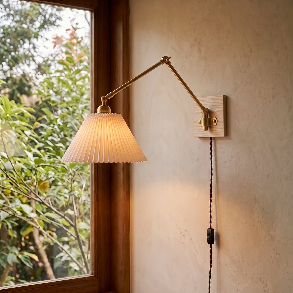 Plug-in wall sconce with a lit pleated shade and brass arm beside a window, warm light on a cream plaster wall