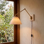 Plug-in wall sconce with a lit pleated shade and brass arm beside a window, warm light on a cream plaster wall