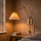 Bedside plug-in swing arm wall sconce with a lit pleated shade on a peach plaster wall above a wooden nightstand