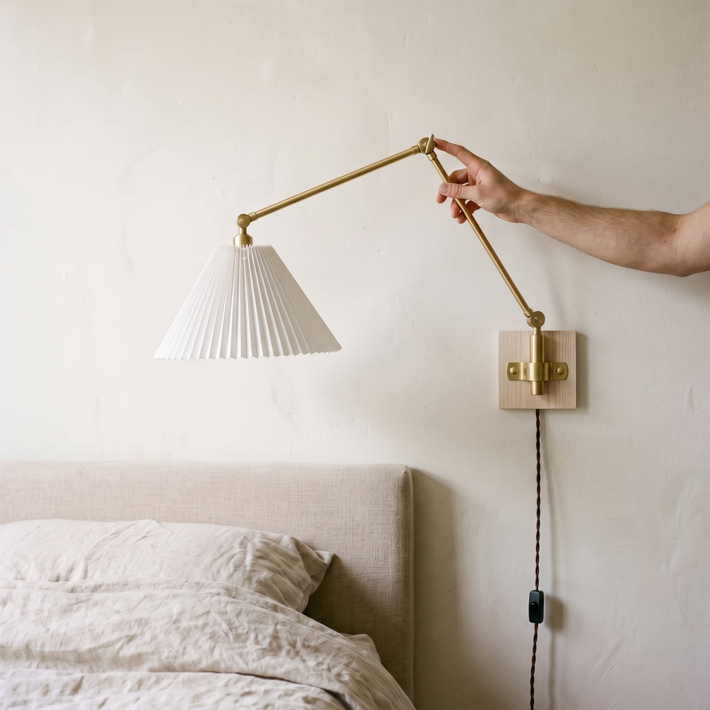 Hand adjusting the folding brass arm of a plug-in swing arm wall sconce with a pleated shade above a bed
