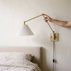 Hand adjusting the folding brass arm of a plug-in swing arm wall sconce with a pleated shade above a bed
