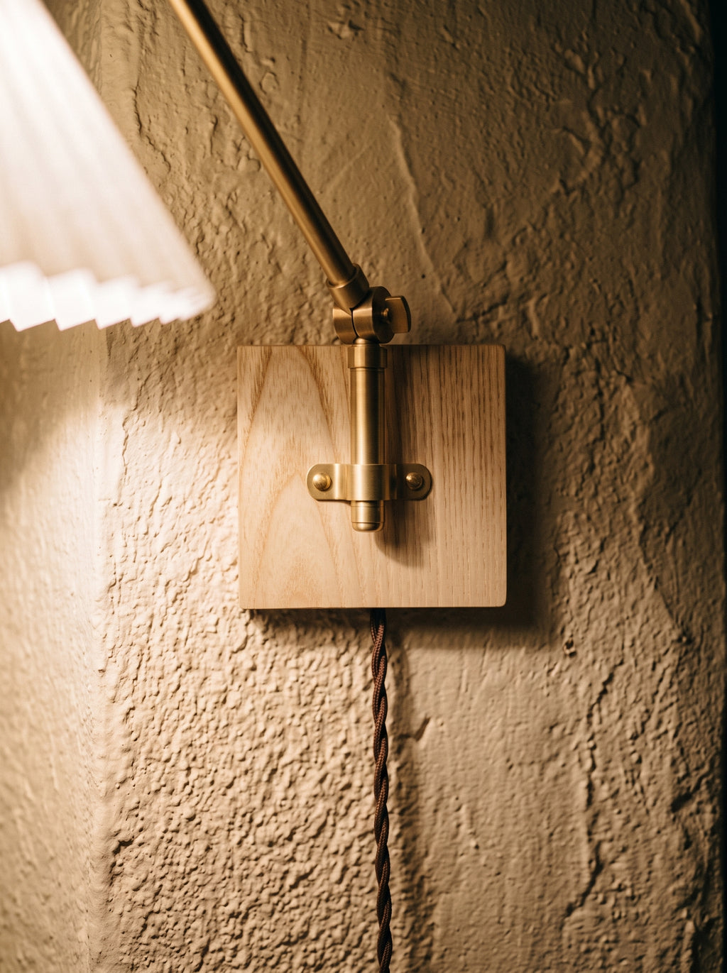 Ash wood wall plate and brass hardware of a plug-in swing arm wall sconce on textured plaster, with the cord running down