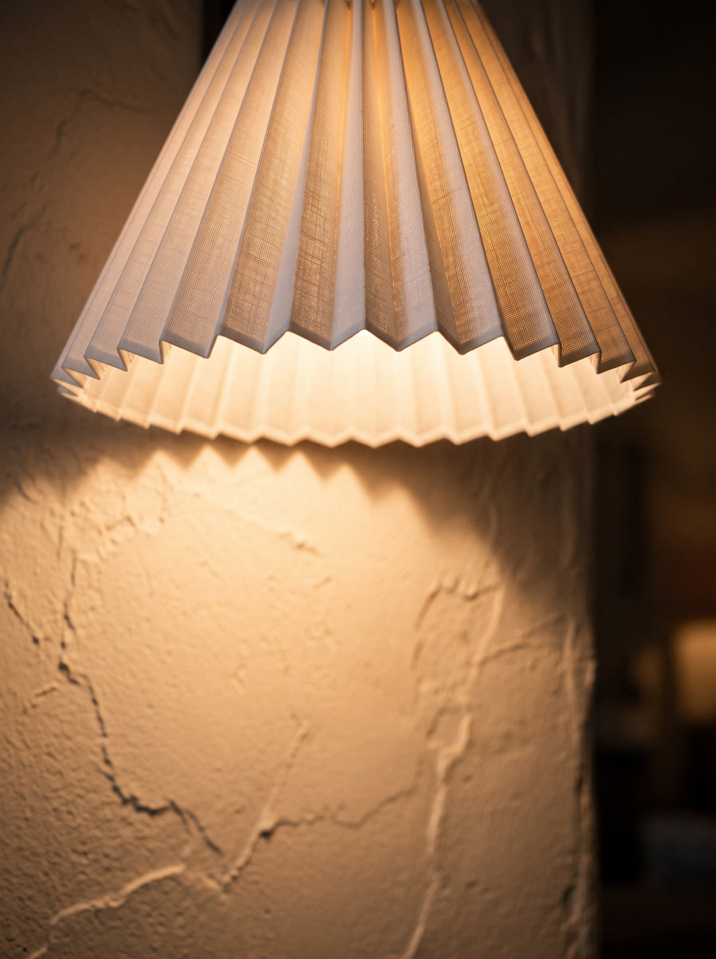 Close view of a lit pleated lamp shade on a wall sconce, warm light spreading over textured plaster
