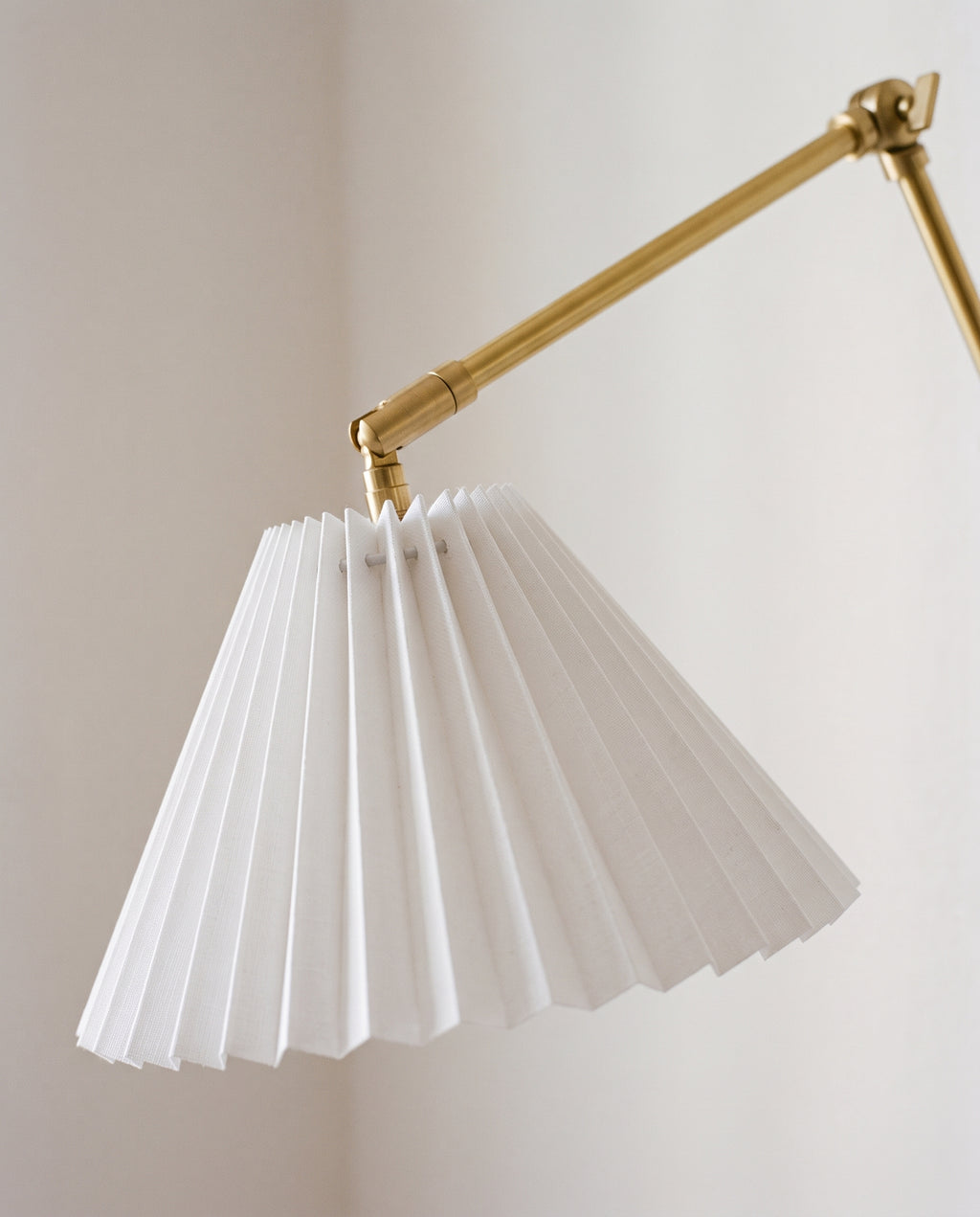 Close view of a white pleated shade on a brass swing arm wall sconce, unlit against a pale wall