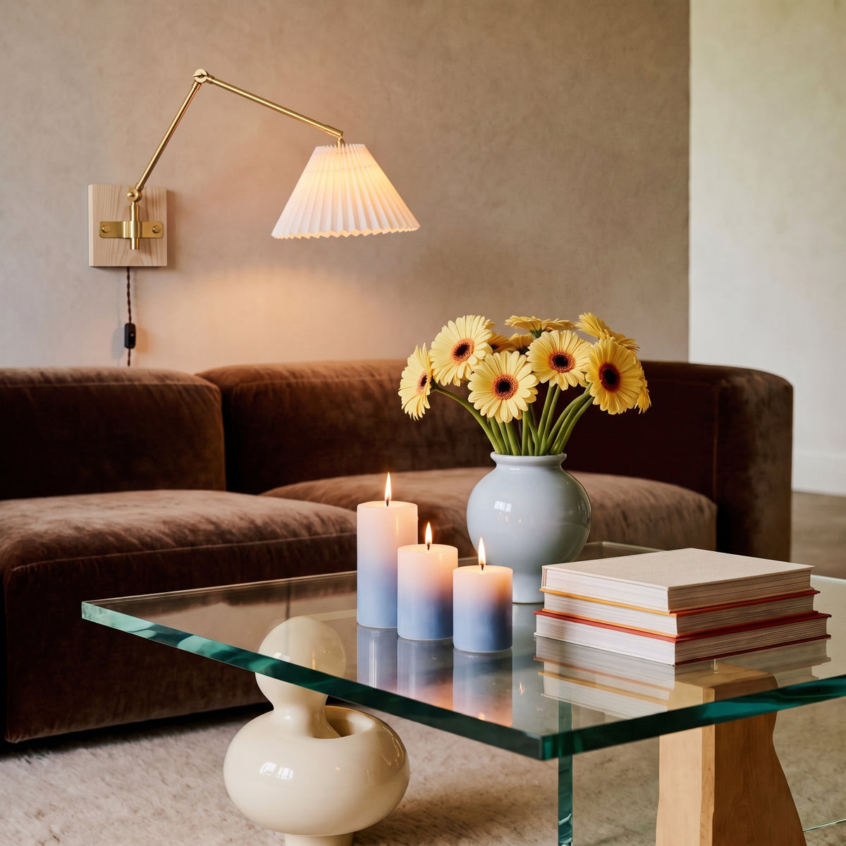 Plug-in swing arm wall sconce with a pleated shade above a brown sofa and a glass coffee table with yellow gerberas and candles