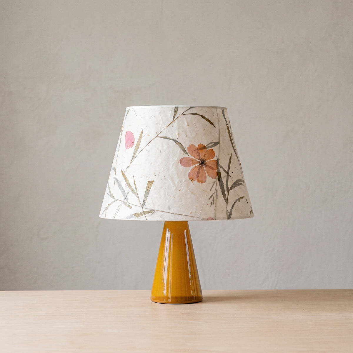 Amber glazed ceramic cone table lamp with a large pressed flower and grass paper shade on a plain backdrop