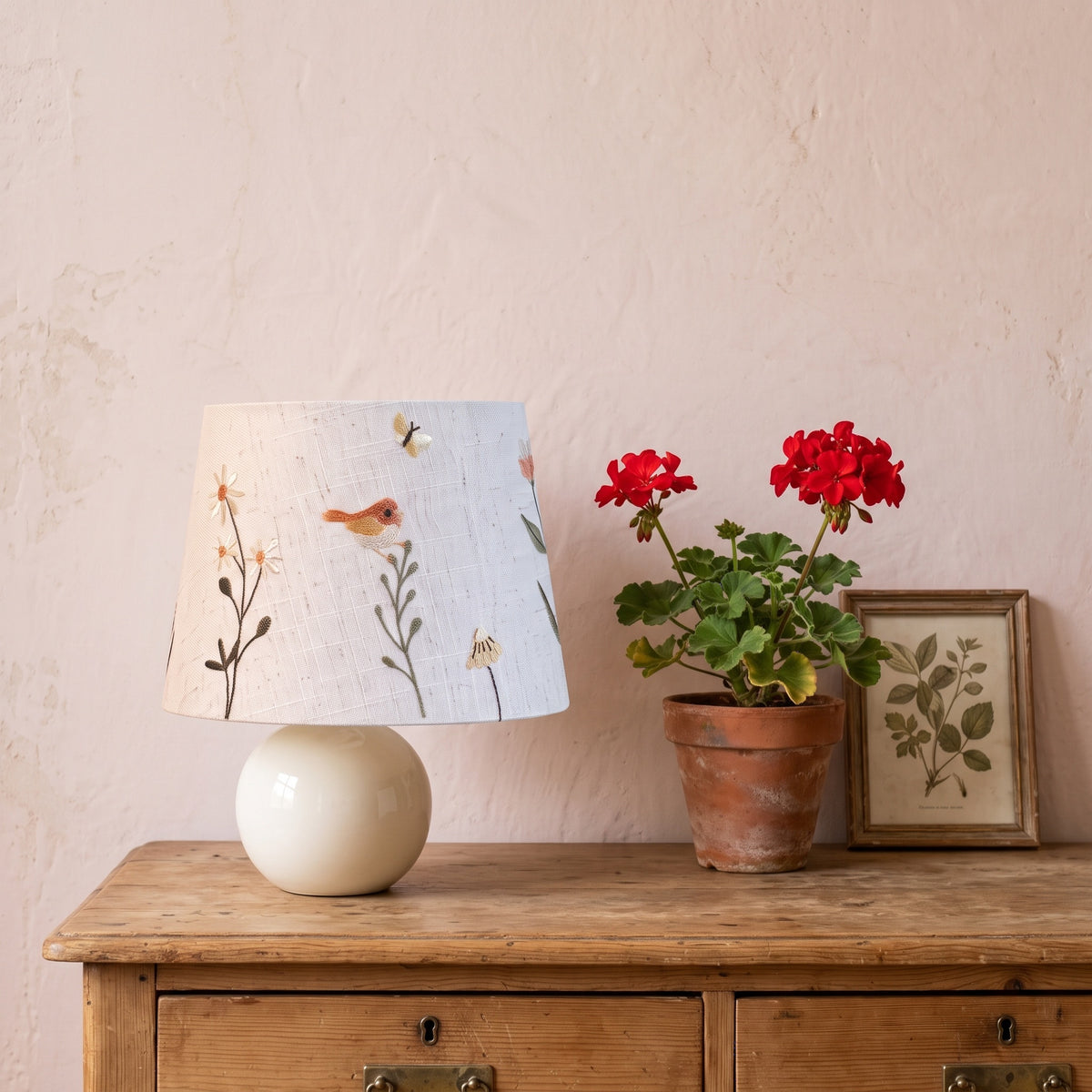 Bird and daisy embroidered table lamp on a pine chest of drawers beside red geraniums and a framed botanical print