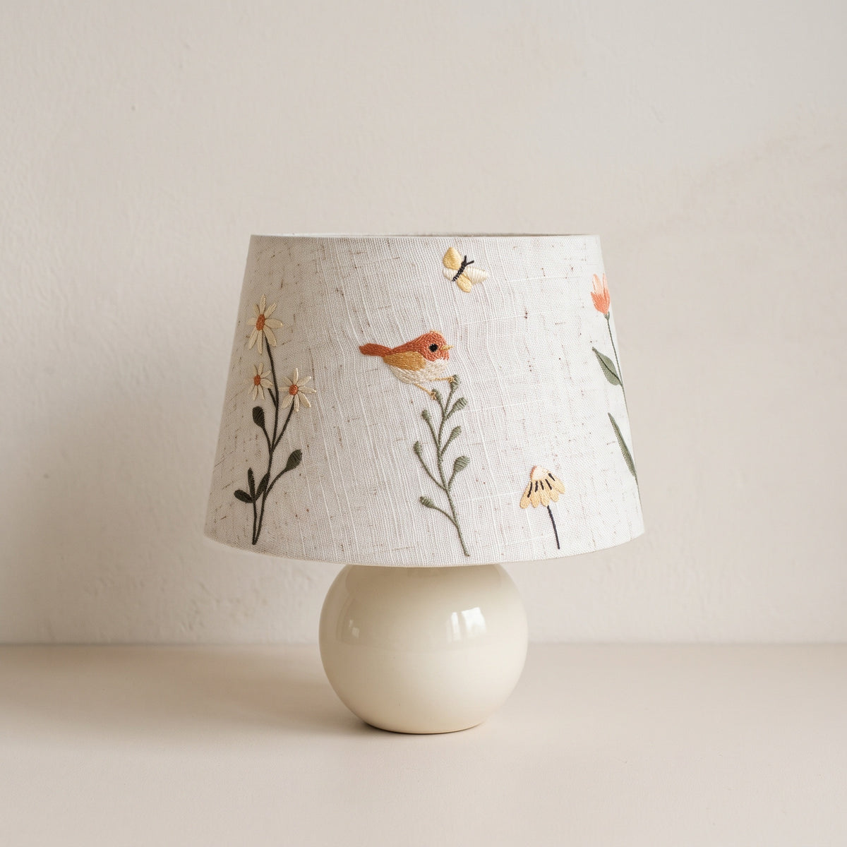 Cream ceramic ball table lamp with a linen-look shade embroidered with a small bird, daisies and a butterfly, on a plain plaster backdrop