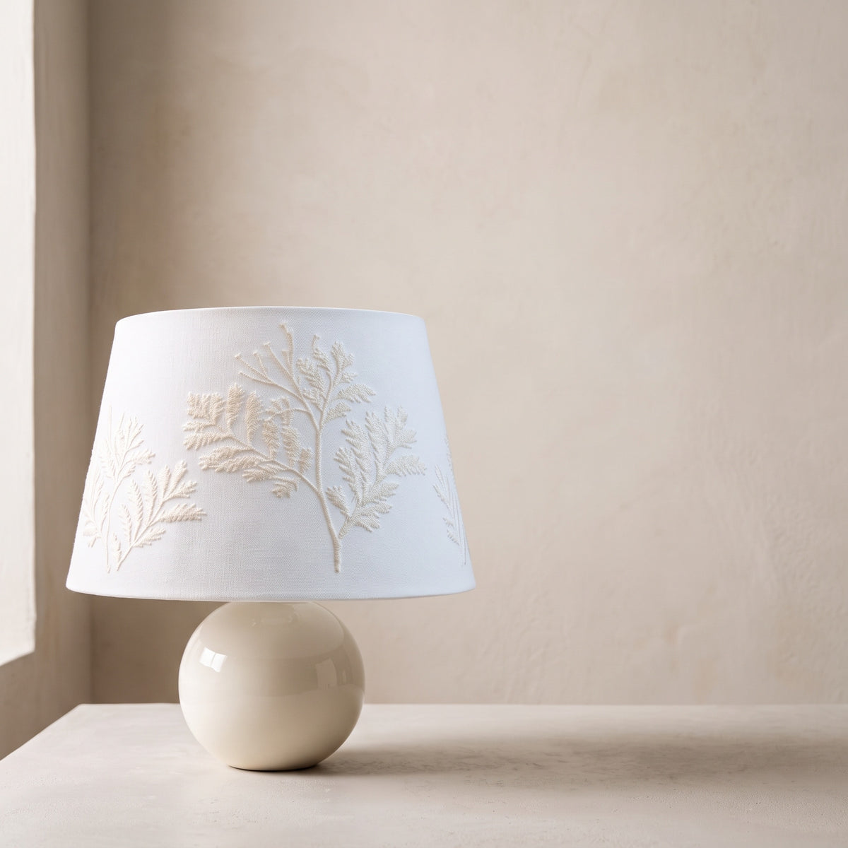 Cream ceramic ball table lamp with a white fern-embroidered drum shade on a plain plaster backdrop