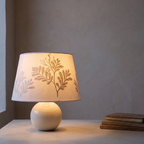 Fern-embroidered table lamp switched on, the shade glowing warm against a grey wall beside a stack of books