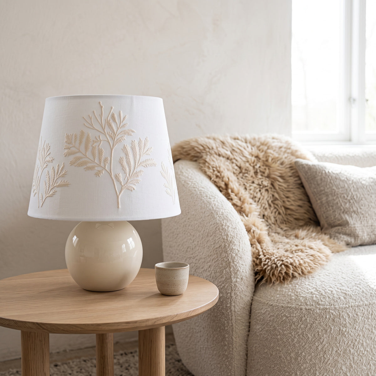 Fern-embroidered table lamp on a round oak side table beside a cream boucle sofa and a sheepskin throw