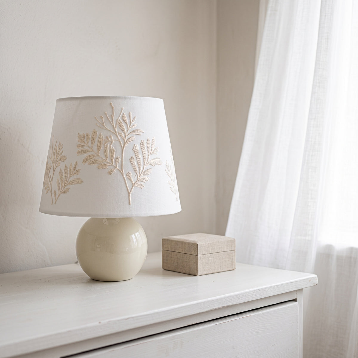 Fern-embroidered table lamp on a white chest of drawers by a sheer curtain, next to a small linen box