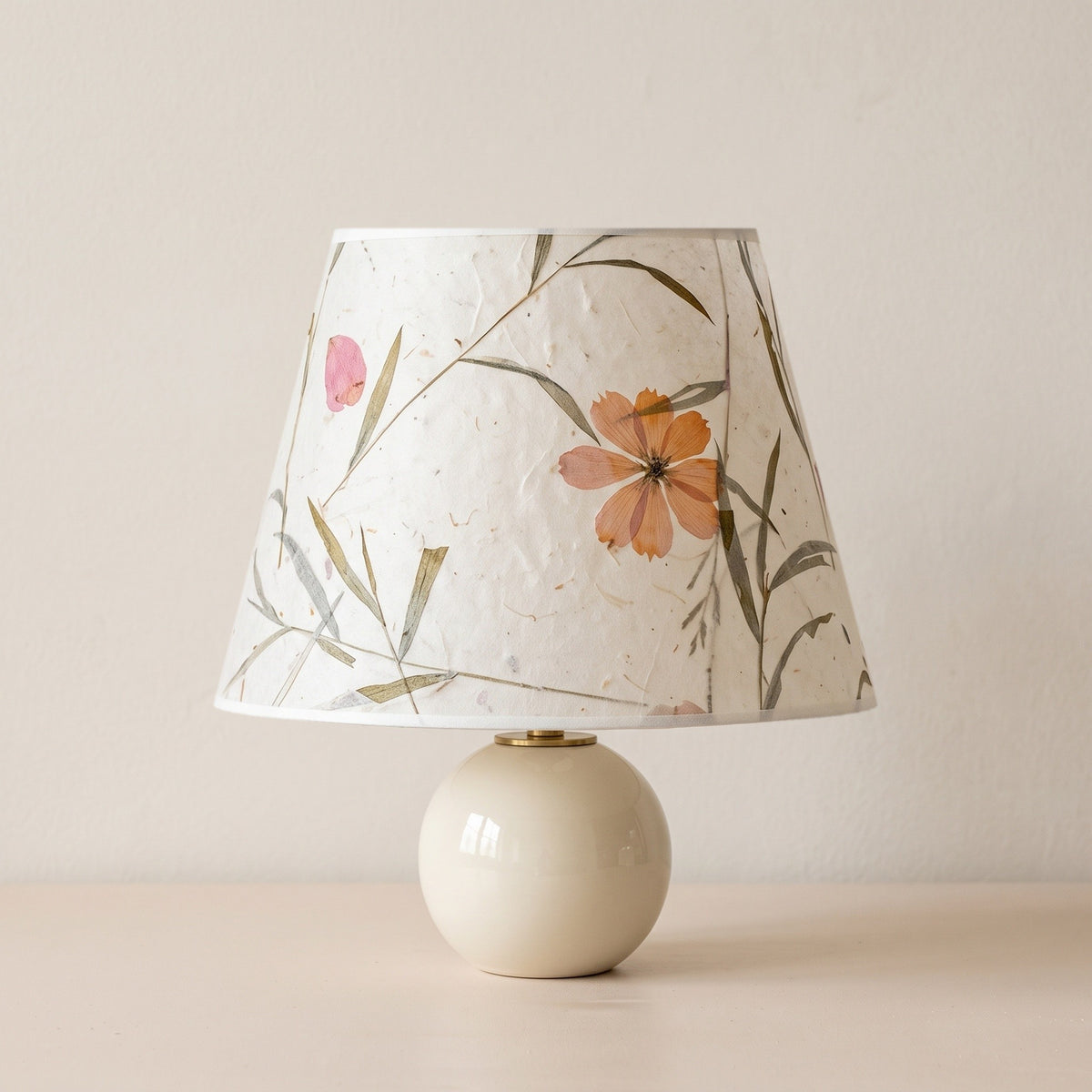 Cream ceramic ball table lamp with a large pressed flower and grass paper shade on a plain plaster backdrop