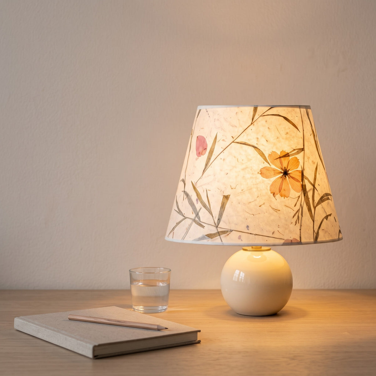 Pressed flower paper lamp switched on at a pale oak desk beside a closed linen notebook and a glass of water