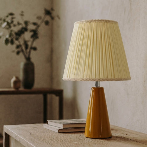 Amber glazed ceramic table lamp with a cream pleated shade on a wooden console table beside two books and a vase of branches