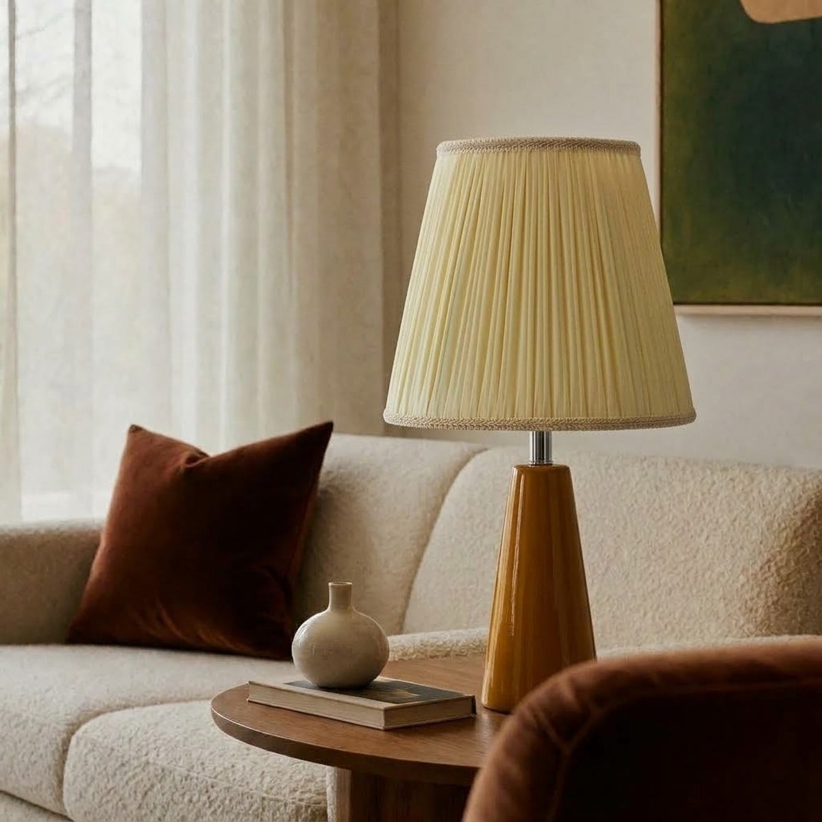 Amber glazed ceramic table lamp with a cream pleated shade on a round wooden side table beside a cream boucle sofa, a rust velvet cushion and a small vase