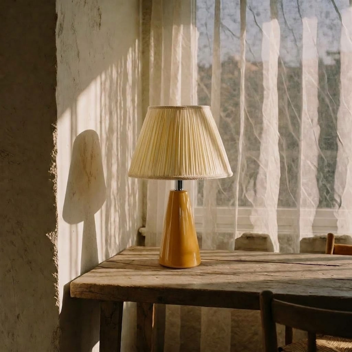 Amber glazed ceramic table lamp with a cream pleated shade on a rustic wooden table beside a sheer curtain and a plaster wall in low sun