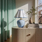 The Blue Wave on a fluted wooden side table beside green curtains, books and a small plant