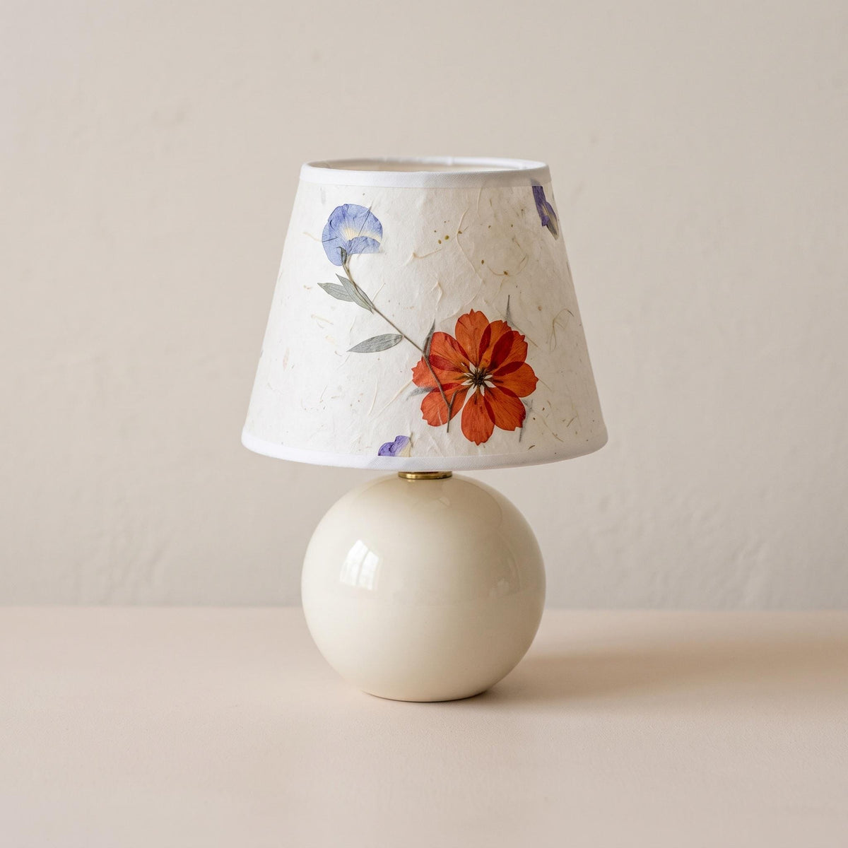 Cream ceramic ball table lamp with a small pressed flower paper shade on a plain backdrop