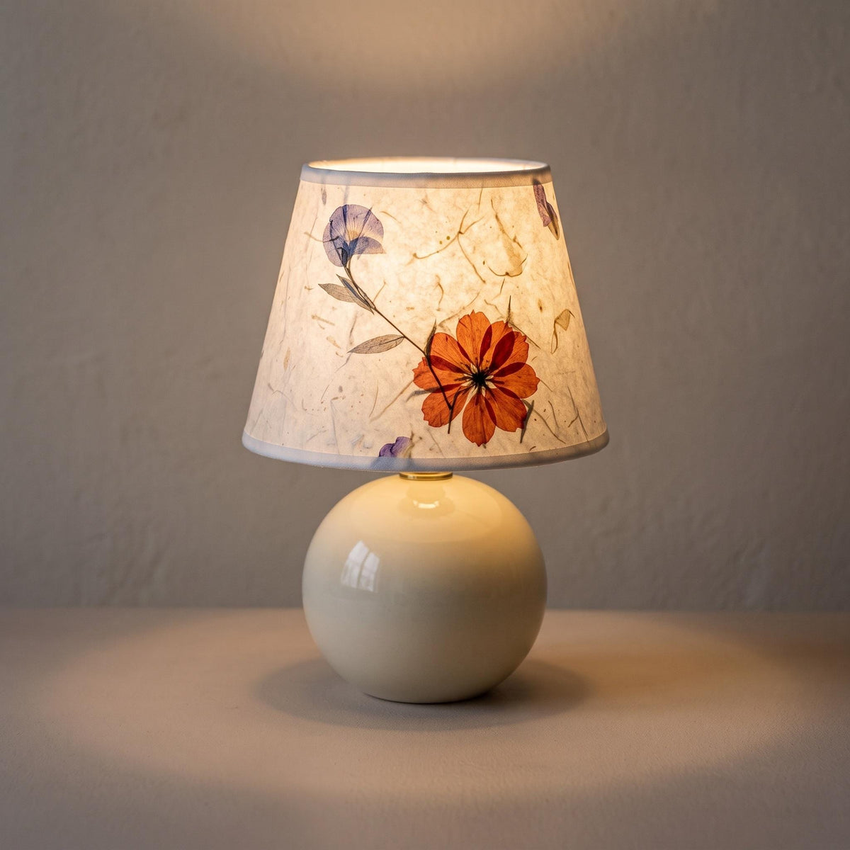 Cream ceramic ball table lamp switched on, the small pressed flower paper shade glowing warm with a red-orange flower and a violet-blue blossom